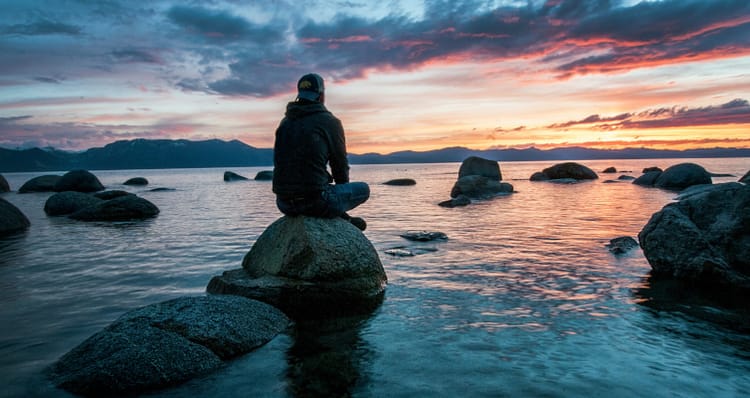 A person sits on a rock in the water, facing away from the camera, and overlooking a lake at sunset. The sky is a mix of blue, orange, and pink hues.